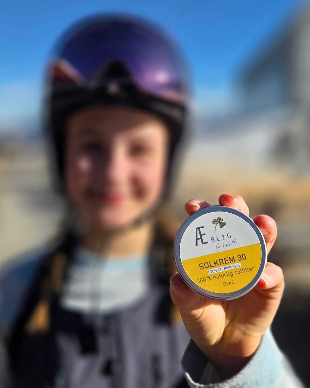 jente med skihjelm holder ærlig by walle solkrem mot kamera, blå himmel